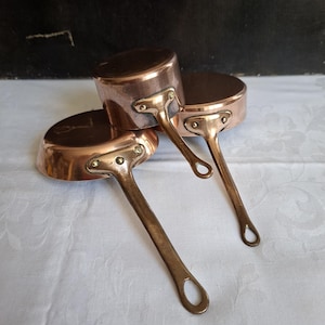 May include: Three copper saucepans with polished metallic surfaces and brass handles. Each pan features a rounded body and a long, slender handle attached with three visible rivets. The pans are stacked together, showcasing their warm, reflective finish.