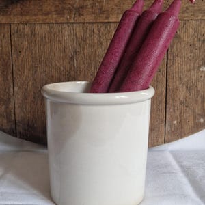 May include: A white ceramic container holding several long, deep purple candles. The candles have a textured appearance and are tied with a red and white string. The container sits on a white cloth, with a wooden background.