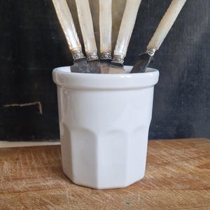 Puede incluir: Un recipiente de cerámica blanca que contiene cinco cuchillos pequeños. Los cuchillos tienen mangos de nácar y detalles metálicos plateados. El recipiente está sobre una superficie de madera.