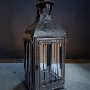 May include: A rectangular metal lantern with a dark, weathered, and rusted finish. It features vertical metal bars protecting glass panels on each side, a hinged door with a small latch, and a circular metal handle at the top for hanging.