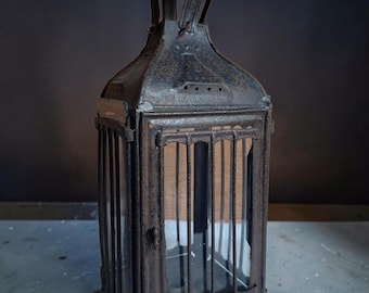 May include: A rectangular metal lantern with a dark, weathered, and rusted finish. It features vertical metal bars protecting glass panels on each side, a hinged door with a small latch, and a circular metal handle at the top for hanging.