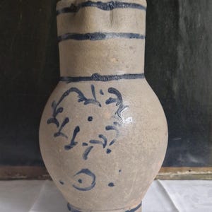 May include: A stoneware jug with a rounded body and a slender neck. The jug is a pale grey colour with dark blue bands and floral patterns. The jug is likely an antique or vintage piece.