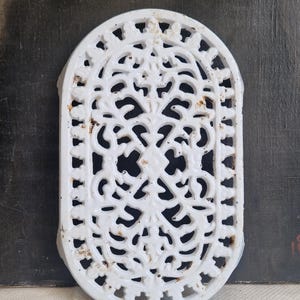 May include: White cast iron trivet with an oval shape and intricate openwork design. The trivet features a detailed pattern of swirling lines and floral motifs. It has a slightly aged appearance with some rust spots.