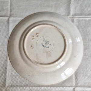 May include: A white ceramic plate with a slightly off-white rim. The underside features the text "Vegear Longchamp" in a circular design, along with the numbers "40" and "56". The plate has a vintage aesthetic.