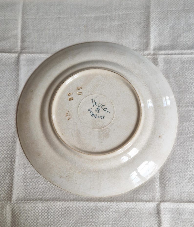 May include: A white ceramic plate with a slightly off-white rim. The underside features the text "Vegear Longchamp" in a circular design, along with the numbers "40" and "56". The plate has a vintage aesthetic.