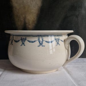 Pode incluir: Bacia de cerâmica creme com um desenho de fita e flores azuis ao redor da borda. A bacia tem um corpo arredondado, uma alça e uma borda larga e plana. O fundo é uma imagem escura e desfocada.