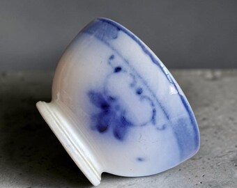 May include: A small white ceramic bowl featuring a hand-painted blue floral pattern around the exterior rim. The bowl has a rounded body and a raised circular base, with the blue pigment appearing slightly blurred and soft against the white glaze.