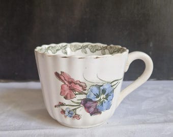 May include: A white teacup with a floral design featuring pink, blue, and purple flowers. The rim has a green leaf pattern. The cup has a handle and a fluted design.