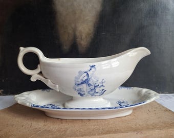 May include: A cream-coloured ceramic gravy boat with a matching oval underplate. The gravy boat features a blue transferware illustration of a figure and a circular handle. The underplate has a blue patterned border and a scalloped edge design.