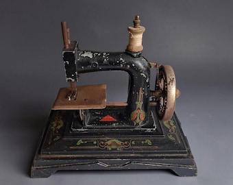 May include: A vintage black cast iron sewing machine with a rectangular base, featuring worn paint, decorative floral decals in red and green, a metal hand wheel, a needle bar, and a wooden spool of thread mounted on the top arm.