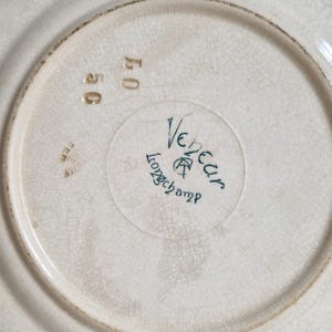 May include: Off-white ceramic plate with a cracked glaze effect. The plate's underside features the words "Vedeur Longchamp" in teal script, along with the numbers "56" and "40" in gold. A vintage, antique aesthetic.