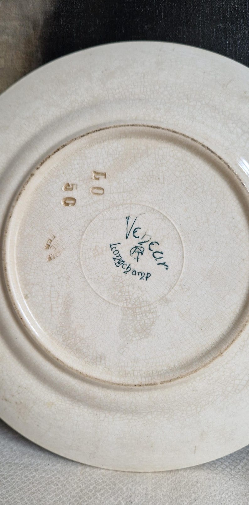 May include: Off-white ceramic plate with a cracked glaze effect. The plate's underside features the words "Vedeur Longchamp" in teal script, along with the numbers "56" and "40" in gold. A vintage, antique aesthetic.
