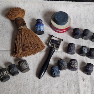 Pode incluir: Uma variedade de artigos de escritório vintage, incluindo uma pequena escova, um frasco de tinta, uma almofada de tinta e vários selos cilíndricos decorativos. Os itens estão dispostos em uma superfície branca.