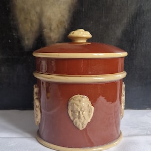 Puede incluir: Un tarro de cerámica con tapa, con un esmalte marrón rojizo y detalles crema. El tarro está adornado con esculturas en relieve de rostros. La tapa tiene un pomo decorativo. Una pieza clásica para la cocina o la decoración del hogar.