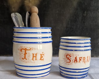 May include: Two white ceramic canisters with blue stripes and orange lettering. One reads "THE" and contains utensils and a wooden figure. The other reads "SAFRAN". Decorative kitchen storage.