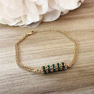 May include: A gold-colored bracelet with a double chain design. The bracelet features a central cylindrical element adorned with multiple emerald-green gemstones. The bracelet is displayed on a light-colored wooden surface.