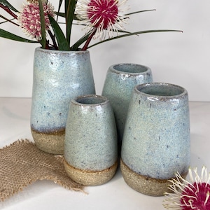 May include: A set of four ceramic vases with a speckled light blue glaze on the upper portion and a textured, sandy-coloured base. The vases are cylindrical with slightly tapered openings and contain stems with spiky pink and white flowers.
