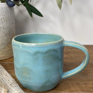 May include: A handmade ceramic mug with a textured, wavy surface finished in a vibrant turquoise blue glaze. The mug features a rounded body, a wide opening, and a curved handle extending from the side, showcasing a glossy, uneven finish.