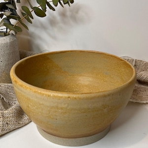 May include: A round ceramic bowl with a warm, golden-yellow glazed interior and exterior. The bowl features a smooth, curved rim and a distinct, unglazed light grey base with a speckled texture, showcasing a handcrafted, functional design.