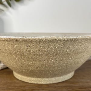 May include: A wide, shallow ceramic bowl with a speckled, sandy-beige finish. The exterior surface features a rough, textured appearance with visible dark flecks throughout the clay body, leading down to a circular, slightly raised footed base.
