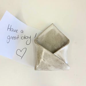 May include: A ceramic envelope-shaped holder with a speckled off-white glaze. A white note is tucked inside, featuring the handwritten black ink text "Have a great day!" followed by a hand-drawn heart symbol on the paper surface.