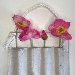 May include: A handmade ceramic wall vase with a white macramé handle, holding pink poppies with yellow centres. The vase has a textured, ridged design and is a neutral off-white colour.
