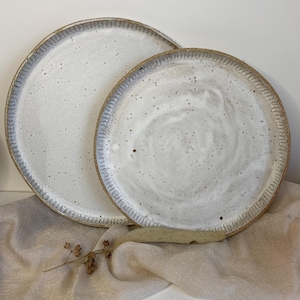 May include: Two handmade ceramic plates. The plates are white with speckled detailing and a textured rim. The rims have a blue-grey accent. The plates are of different sizes and are displayed on a beige cloth.