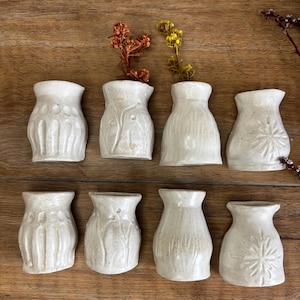 May include: Eight small, off-white ceramic wall vases arranged in two rows. Each vase features a unique embossed pattern, including vertical lines, floral stems, and starburst designs. Two vases contain small dried orange and yellow flowers.