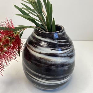 May include: A small, rounded ceramic vase with a glossy finish featuring a swirling marbled pattern of black and white. A sprig of green leaves and a bright red bottlebrush flower extend from the opening of the vase, adding a natural element.