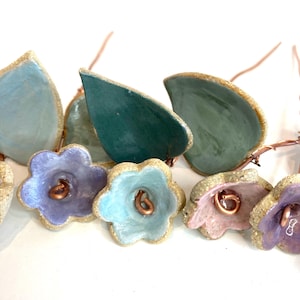 May include: Close-up of handmade ceramic flowers and leaves in pastel colours. The flowers have copper wire centres and gold-coloured edges. The leaves are in shades of blue and green.