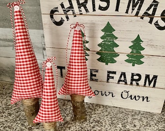 May include: Three red and white gingham fabric Christmas trees with white buttons on top, each with a brown twig base. The trees are arranged on a speckled countertop in front of a white sign that reads "Christmas Tree Farm Your Own".