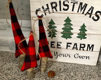 May include: Three small Christmas trees made of red and black plaid fabric with a burlap bow on each. The trees are attached to wooden branches and are sitting on a granite countertop. A wooden sign behind the trees reads "Christmas Tree Farm Your Own".