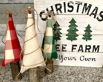 May include: Three handmade Christmas tree decorations. The trees are made of fabric with a plaid pattern in red and white, green and white, and white. Each tree is wrapped with twine and has a wooden base. The trees are displayed in front of a white sign that reads "Christmas Tree Farm Your Own".
