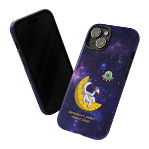 Puede incluir: Una funda de teléfono con temática espacial. La funda presenta un fondo de galaxia púrpura con un astronauta en una luna creciente, un cohete y un OVNI. El texto "WRONG PLANET RIGHT VIBE" está impreso en la funda.