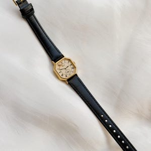 May include: A gold-toned wristwatch with a black leather band. The watch face is octagonal with Roman numerals and a small subdial. The band has a gold buckle. The watch is displayed on a white, textured surface.