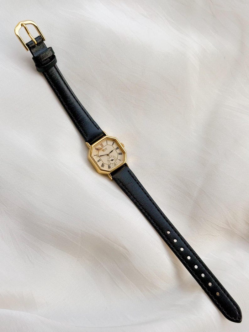 May include: A gold-toned wristwatch with a black leather band. The watch face is octagonal with Roman numerals and a small subdial. The band has a gold buckle. The watch is displayed on a white, textured surface.