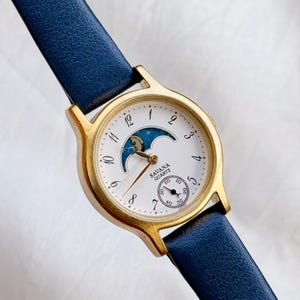 May include: A gold-coloured Savana Quartz watch with a white face, featuring a moon phase design and a small seconds dial. The watch has a navy blue leather strap.