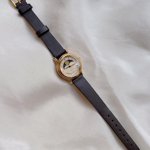 May include: A gold-toned wristwatch with a black leather band. The watch face features a moon phase design with a crescent moon and stars. The watch has a classic, elegant style.