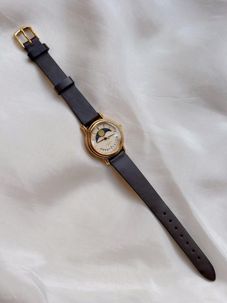 May include: A gold-toned wristwatch with a black leather band. The watch face features a moon phase design with a crescent moon and stars. The watch has a classic, elegant style.