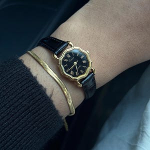 May include: A gold wristwatch with a black face and black leather strap. The watch features Roman numeral hour markers and gold hands. A gold bracelet is also visible.