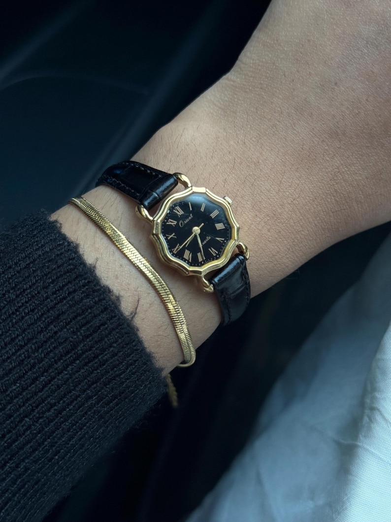May include: A gold wristwatch with a black face and black leather strap. The watch features Roman numeral hour markers and gold hands. A gold bracelet is also visible.