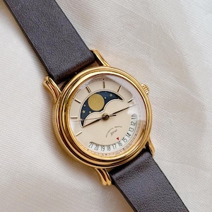 May include: A gold-toned wristwatch with a cream-colored face and a dark brown leather band. The watch face features a moon phase indicator and a date display. The watch has the text "SENTIMENTAL GRAFFITI YOU" on the face.
