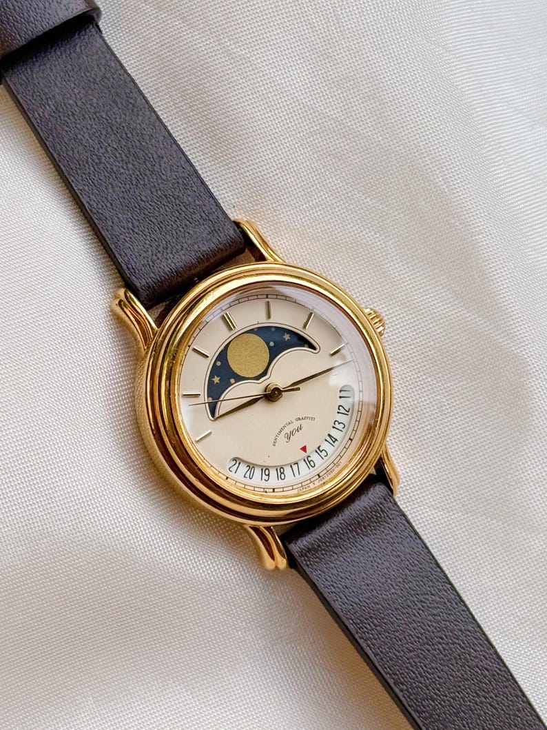 May include: A gold-toned wristwatch with a cream-colored face and a dark brown leather band. The watch face features a moon phase indicator and a date display. The watch has the text "SENTIMENTAL GRAFFITI YOU" on the face.