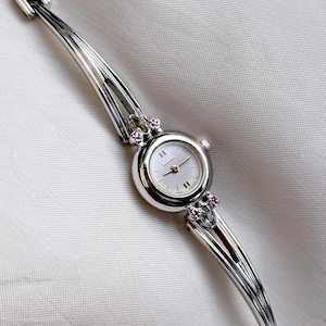 May include: A silver-toned wristwatch with a white face and delicate silver band. The watch face has small silver markers and hands. The band features an intricate design with decorative accents, including small pink gemstones. The watch is displayed on a white surface.
