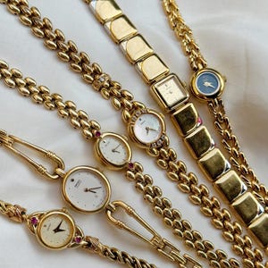 Può includere: Una collezione di orologi da polso vintage color oro con vari stili di cinturino. I quadranti degli orologi sono rotondi, quadrati e rettangolari, con quadranti bianchi o blu scuro. Alcuni orologi presentano pietre preziose e maglie di catena intricate.
