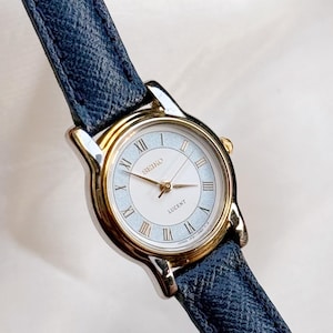 Könnte beinhalten: Eine Seiko Lucent Armbanduhr mit goldfarbener Lünette und hellblauem Zifferblatt. Die Uhr hat römische Ziffern und goldene Zeiger. Sie hat ein marineblaues Lederarmband.