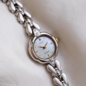 May include: A Seiko Exceline wristwatch featuring a round mother-of-pearl dial with gold-tone hands and markers. The watch case is silver-tone with a gold-tone bezel and two small clear stones at the top and bottom. The bracelet has a matte silver-tone link design.