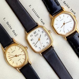 May include: Three vintage gold-toned wristwatches with black leather bands. The watches have white or cream-colored faces with black numerals and hands. The first watch is a Seiko Oval, the second is a Citizen Tank, and the third is a Seiko Tank.