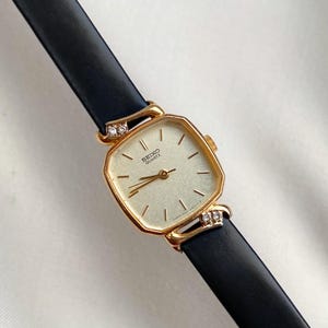 Puede incluir: Un reloj Seiko Quartz vintage con una esfera cuadrada dorada y una correa de cuero negro. El reloj presenta pequeños detalles de diamantes en las fijaciones de la correa. La esfera del reloj tiene un fondo claro con agujas y marcadores negros.