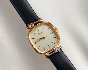 Puede incluir: Un reloj Seiko Quartz vintage con una esfera cuadrada dorada y una correa de cuero negro. El reloj presenta pequeños detalles de diamantes en las fijaciones de la correa. La esfera del reloj tiene un fondo claro con agujas y marcadores negros.